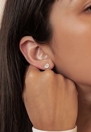 Silberne Ohrstecker mit einem Cluster kleiner klarer Steine, die eine runde Form zeigen. Nahaufnahme von Ohr und Hand, die das Ohrläppchen hält.