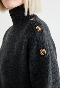 Suéter de punto gris oscuro con un alto cuello acanalado, que presenta tres grandes botones brillantes en el hombro y un acabado suave y texturizado.