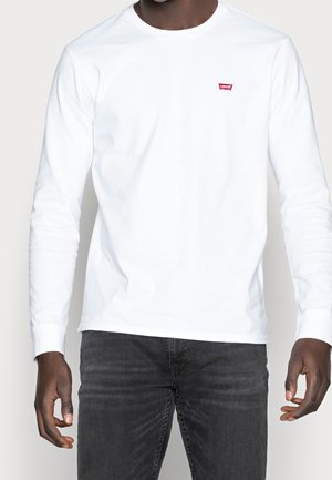 Mand iført simpel hvid langærmet skjorte med lille rød Levi's-logo på brystet, kombineret med falmede sorte jeans.