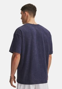 Mann mit kurzen Haaren und Ohrstecker, der ein verwaschenes navyblaues, übergroßes T-Shirt trägt, mit dem Rücken zu einer schlichten weißen Wand.