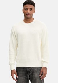 Maglione lavorato a maglia color crema con scollo rotondo, polsini e orlo a coste. Presenta un piccolo logo sul petto. Indossato con jeans scuri.