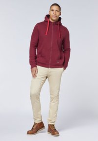 Burgunderfarbener Reißverschluss-Hoodie mit roten Kordeln, zwei Fronttaschen, kombiniert mit beigen Hosen und braunen Schnürstiefeln. Weiche Textur, lässiges Design.