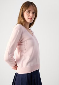 Šviesiai rožinė v-kaklo megztinis ilgomis rankovėmis, su dekoratyvinėmis skylutėmis kairėje pusėje. Minkšta tekstūra ir prigludusi, raukšlėta apykaklė ties juosmeniu.