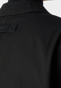 Vue en gros plan de la partie supérieure arrière d'une veste noire avec une boucle en tissu cousue portant le nom de la marque "HUGO."