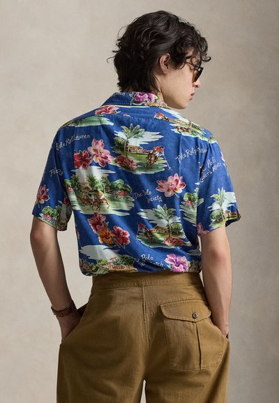 Homme portant une chemise bleu à manches courtes avec des motifs floraux et de joueur de polo, associée à un pantalon marron, tourné de dos les mains dans les poches.
