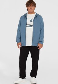 Blue zip-up hoodie with drawstring, white graphic t-shirt, black pants, and white sneakers. Smooth fabric, casual fit, minimal accents.