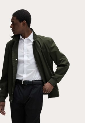 Mand, der drejer til venstre iført en olivengrøn jakke over en hvid skjorte med knapper, sorte bukser og sort bælte.