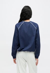 Tengerészkék könnyű dzseki hosszú ujjakkal és fehér díszítő csíkkal a varrások mentén. Laza szabású, kerek nyakkivágással és elasztikus mandzsettákkal.