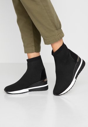 Person wearing black knit sock-style ankle sneakers with white and black wedge soles, paired with olive green cropped pants.