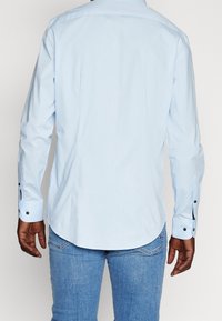 Mężczyzna w jasnoniebieskiej koszuli z długim rękawem zapinanej na guziki i niebieskich dżinsach, przedstawiony z tyłu na jednolitym tle.