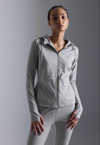 Lys grå idrettsjakke med glidelås foran, hette og hull for tomlene, kombinert med matchende grå leggings. Glatt, elastisk stoff.