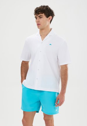 Mann trägt ein weißes kurzärmeliges Hemd mit Knöpfen und leuchtend blaue Shorts, steht mit Händen in den Taschen vor einem einfarbigen Hintergrund.