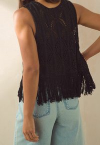 Schwarzes gehäkeltes Top mit lockerer Passform, das über ein Rautenmuster und Fransenverzierungen am Saum verfügt. Kombiniert mit hellblauen Jeans.