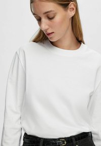 T-shirt bianco a maniche lunghe con scollatura rotonda, realizzato in cotone morbido, indossato con pantaloni neri a vita alta con una cintura strutturata.