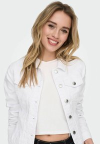 White denim jacket with metal button closures, two chest pockets, and a fitted design. Worn over a light-colored, ribbed top.