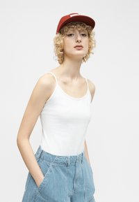 Jeune personne aux cheveux blonds bouclés, portant une casquette rouge, un débardeur blanc et un pantalon bleu rayé, les mains dans les poches, regardant devant elle.