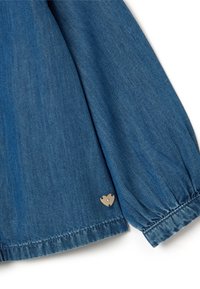 Dettaglio di un indumento in denim con maniche lunghe e arricciate di colore blu. Presenta un piccolo accento metallico a forma di cuore cucito vicino all'orlo.