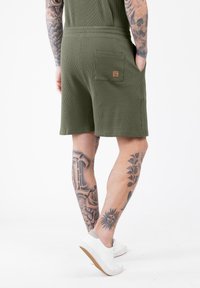 Man met getatoeëerde armen en benen draagt olijfgroene, gestructureerde shorts met een achterzak en witte sneakers, staande tegen een witte achtergrond.