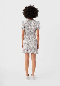 Kvinna med lockigt hår iklädd en vit blommig kortärmad klänning med volangkant och smockad midja, stående med ryggen mot kameran.