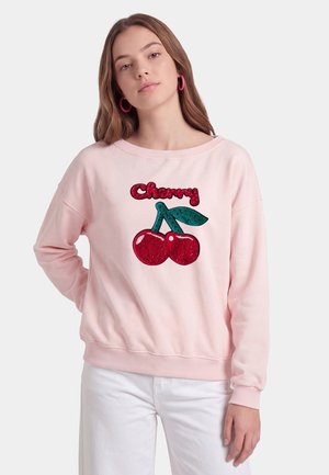 Młoda kobieta o długich brązowych włosach, ubrana w jasnoróżową bluzę z czerwonymi wisienkami i słowem "Cherry,", w połączeniu z białymi spodniami.