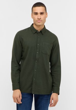 Junger Mann, der ein dunkelgrünes Langarmhemd mit Knöpfen und einer Brusttasche sowie blaue Jeans trägt, im Stehen vor einem schlichten weißen Hintergrund.