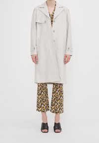 Lys grå trenchcoat med nøglekrave, enkeltstående design og store lommer, lagd over mønstrede bukser og sorte mule-sko.