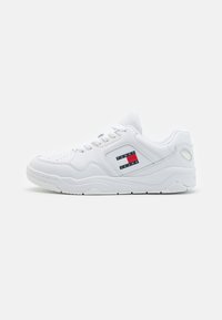 Zapatilla de cuero blanca con puntera perforada, forma redondeada, diseño con cordones y un parche lateral que presenta acentos en rojo y azul marino.
