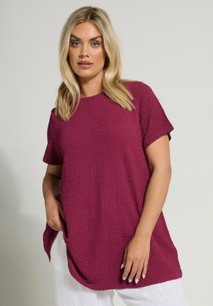 Blondynka w teksturowanej, krótkiej tunice w ciemnoróżowym kolorze i białych spodniach, stojąca na jasnym, jednolitym tle.