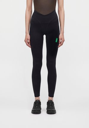 Person iført sorte leggings med høj talje og et grønt mærke på låret samt sorte sportssko, stående foran en ensfarvet lys baggrund.