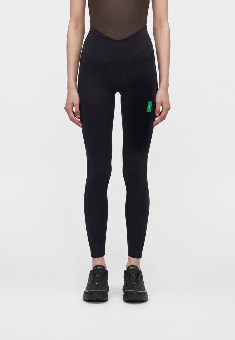 Personne portant des leggings noirs taille haute avec une étiquette verte sur la cuisse et des chaussures de sport noires, debout devant un fond clair uni.
