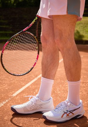 Scarpe da tennis bianche con accenti grigi, parte superiore in rete e loghi neri; abbinate a calzini sportivi bianchi. Una racchetta da tennis è tenuta accanto alle gambe.