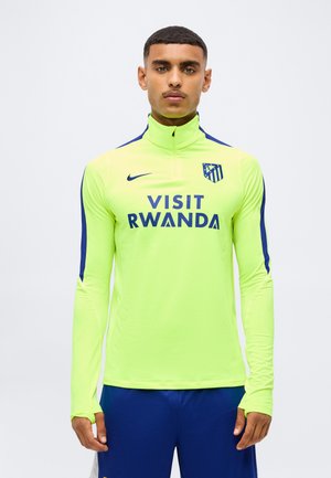 Młody mężczyzna noszący neonowożółty sportowy top z długim rękawem z "Visit Rwanda" i logo drużyny piłkarskiej na piersi oraz niebieskie sportowe spodenki.