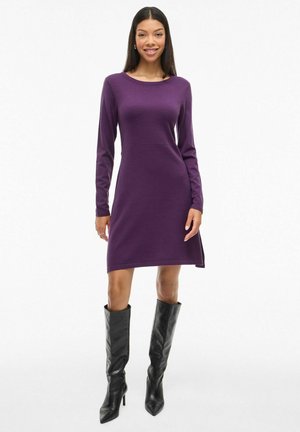 Lila Strickkleid mit langen Ärmeln und Rundhalsausschnitt, mit einem seitlichen Schlitz, kombiniert mit hohen schwarzen Stiefeln. Glatte Textur und enganliegendes Design.