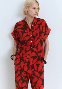 Red and brown short-sleeve shirt with a bold floral pattern, featuring a button front and relaxed fit. Paired with matching trousers.