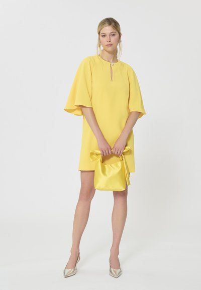 Femme portant une robe jaune ample, tenant un sac à main jaune assorti, et chaussant des talons pointus argentés, debout devant un fond uni.