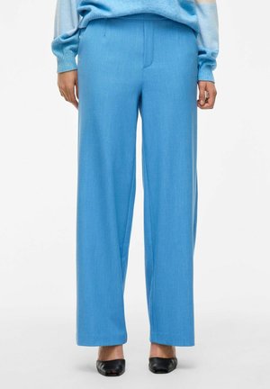 Pantalon large bleu clair en tissu lisse, avec un devant plat et sans motifs visibles. Une paire de talons noirs vient compléter le look.
