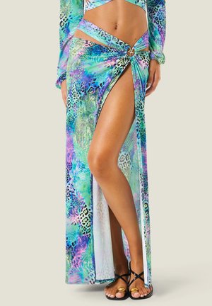 Donna che indossa una gonna lunga colorata con spacco alto e dettaglio anello laterale, abbinata a sandali neri con listini e anelli dorati per le dita dei piedi.