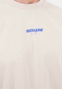 Camiseta de algodón beige con cuello redondo y un logo impreso en azul que dice "SIXTH JUNE PARIS" en el pecho. Textura suave, diseño casual.