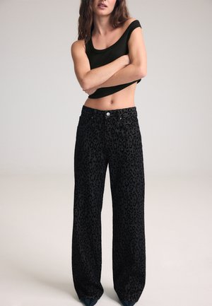 Femme portant un crop top noir épaules dénudées et un pantalon noir taille haute à jambes larges avec un motif léopard subtil.