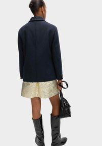 Marineblauer Wollmischmantel mit goldenen Knöpfen, kombiniert mit einem strukturierten goldenen Rock und hohen schwarzen Lacklederstiefeln; hält eine schwarze Handtasche.