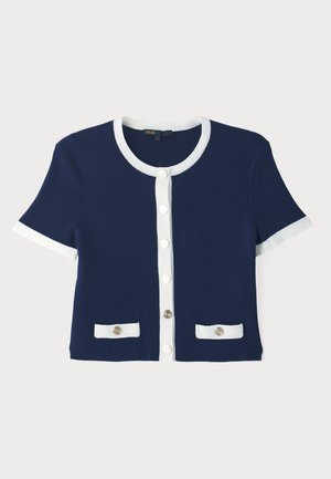 Marineblå ribbstrikket cardigan med hvit kant, korte ermer og frontlukking med knapper. Har to lommer med dekorative knapper.