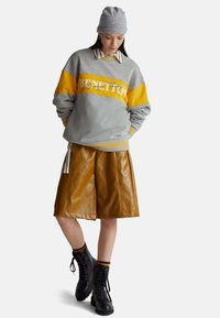 Donna che indossa una felpa grigia e gialla con la scritta "BENETTON", pantaloncini in pelle marrone, berretto grigio, camicia a righe e stivali neri stringati.