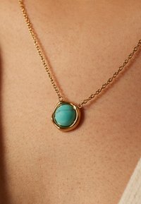 Gouden ketting met een ronde turquoise steen in een onregelmatige metalen setting, met een gladde oppervlakte en aderen.