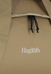 Mochila impermeable marrón con cierres de cordón negros y logotipo bordado en blanco "Haglöfs". Material suave con correas y costuras duraderas.