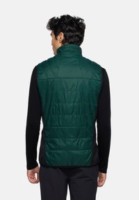 Grüner gepolsterter Weste mit hohem Kragen, die eine gesteppten Struktur und Seitenpaneele aufweist, über einem schwarzen Langarmshirt getragen.