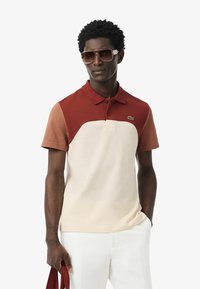 Kortärmad polo-shirt med färgblockdesign i rost, beige och vitt, samt klassisk krage och broderad logotyp på bröstet.