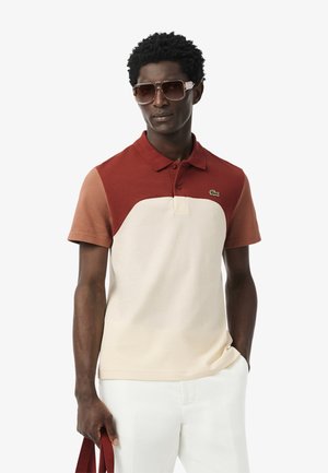 Le polo à manches courtes présente un design en coloris blocs dans des teintes de rouille, beige et blanc, avec un col classique et un logo brodé sur la poitrine.