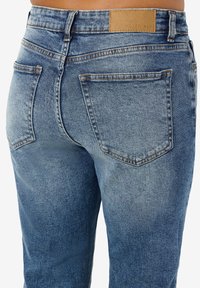 Denimjeans i falmet blå med en glatt tekstur, med sydd rygglommer og en brun lærlapp på midjebåndet.