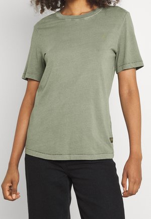 Personne portant un t-shirt uni vert olive à manches courtes et un pantalon noir, montré du cou jusqu’aux hanches, debout, les bras détendus.