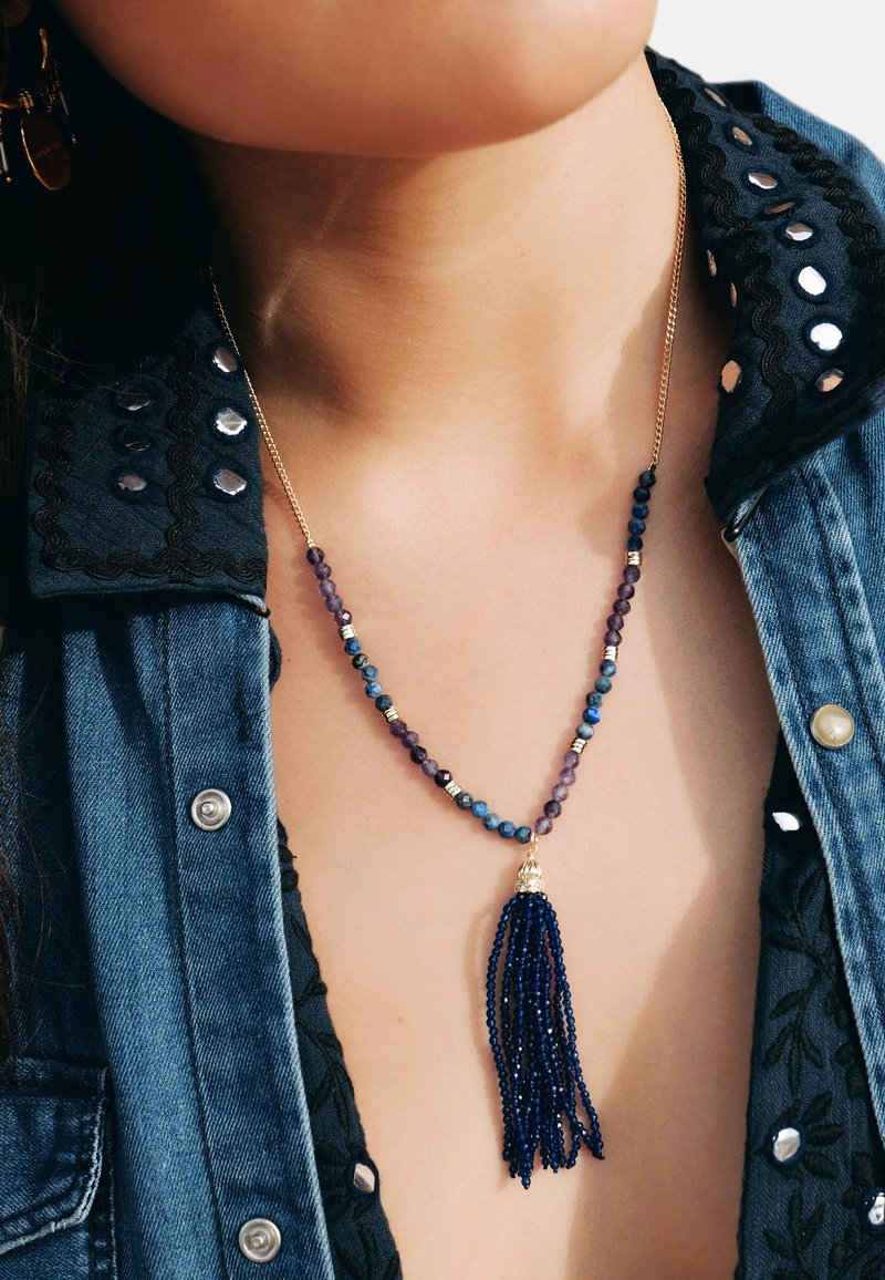 Collier doré avec des perles multicolores et un gland en perles bleues. Présente un mélange de perles rondes et de perles intermédiaires, porté par-dessus une chemise en denim.